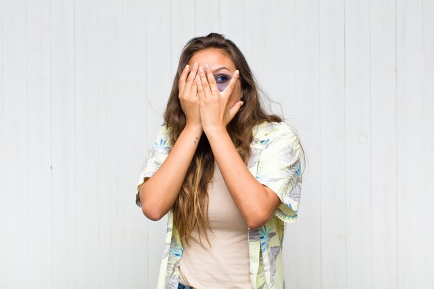 Young pretty woman covering face with hands, peeking between fingers with surprised expression and looking to the side