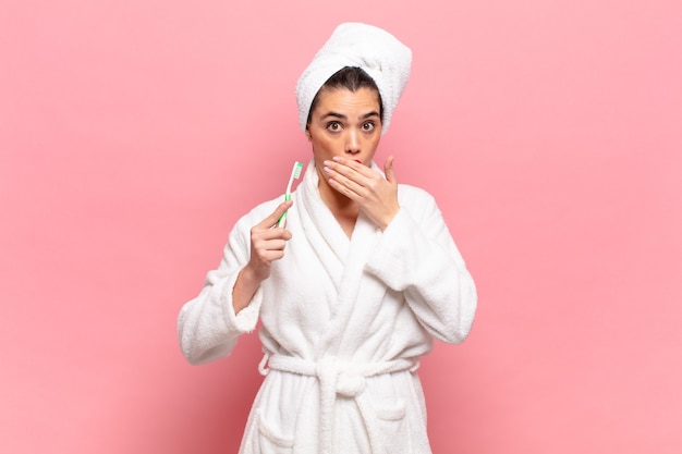 Young pretty woman covering mouth with hands with a shocked, surprised expression, keeping a secret or saying oops. bathrobe and toothbrush concept