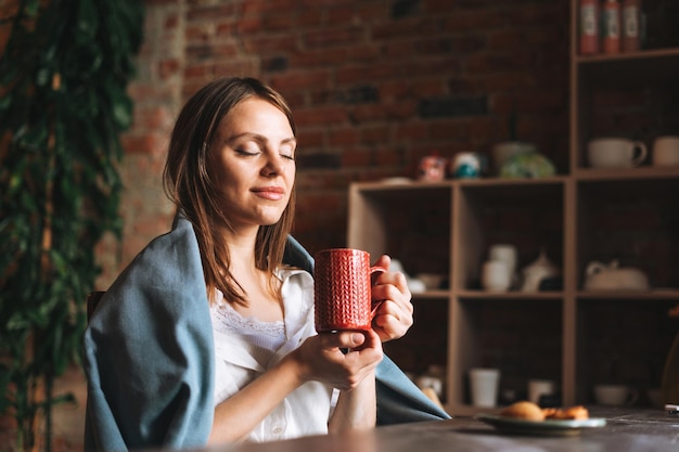 Young pretty woman in cozy grey scarf with mug of tea in hands looks out the window and rests in her studio