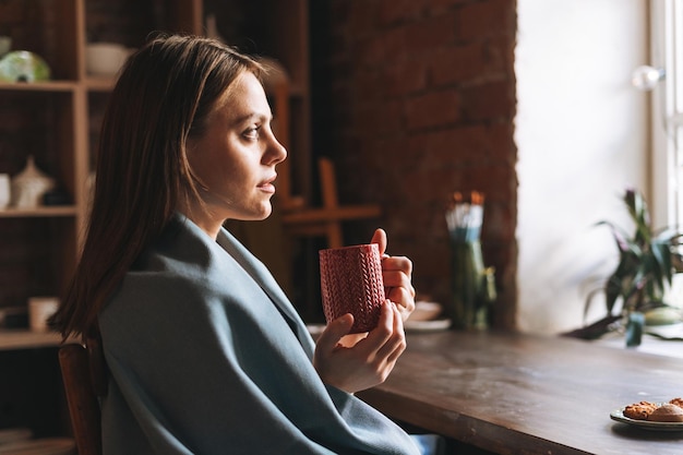 Young pretty woman in cozy grey scarf with mug of tea in hands looks out the window and rests in her studio