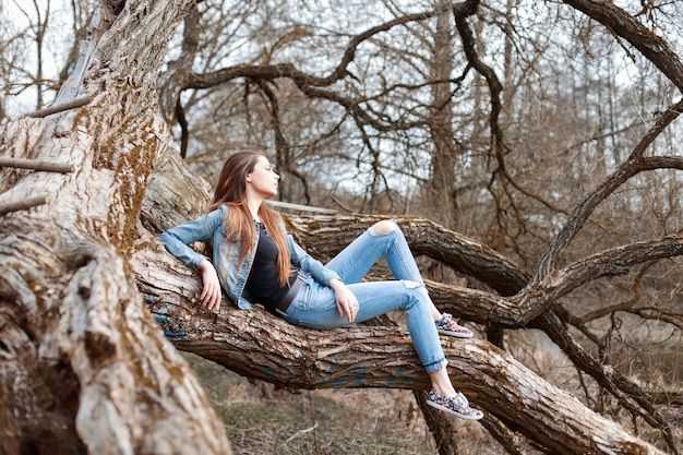 Foto giovane donna graziosa in vestiti del denim che si siedono e che riposa su un grande albero