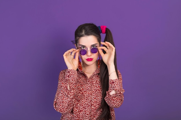 Young pretty woman dressed in 90s style looks with surprise at the camera holding round glasses