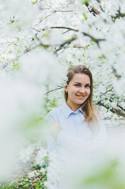 La giovane donna graziosa gode di stare vicino all'albero primaverile in fiore una ragazza che indossa un cappello beige e un abito bianco sorride tra i meli in fiore concetto di stagione primaverile