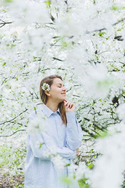 La giovane donna graziosa gode di stare vicino all'albero primaverile in fiore una ragazza che indossa un cappello beige e un abito bianco sorride tra i meli in fiore concetto di stagione primaverile