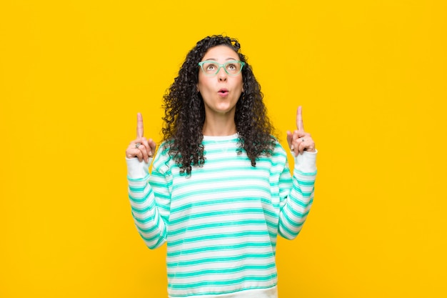 Young pretty woman feeling awed and open mouthed pointing upwards with a shocked and surprised look against orange wall