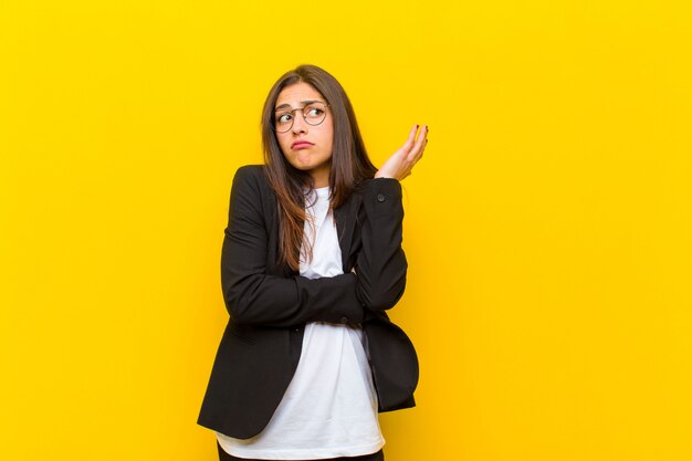 Young pretty woman feeling confused and clueless, wondering about a doubtful explanation or thought against orange wall