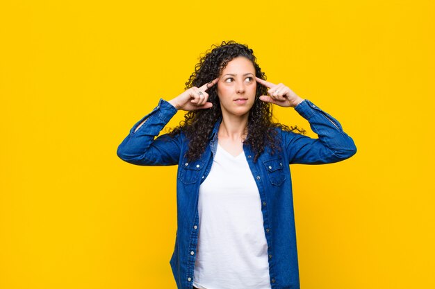 Young pretty woman feeling confused or doubting, concentrating on an idea, thinking hard, looking to copy space on side over orange wall