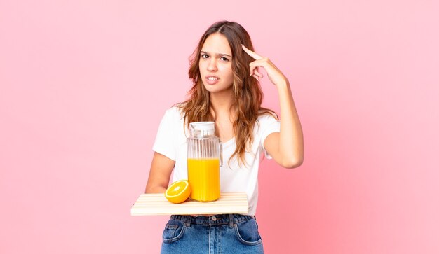 Young pretty woman feeling confused and puzzled, showing you are insane and holding a tray with an orange juice