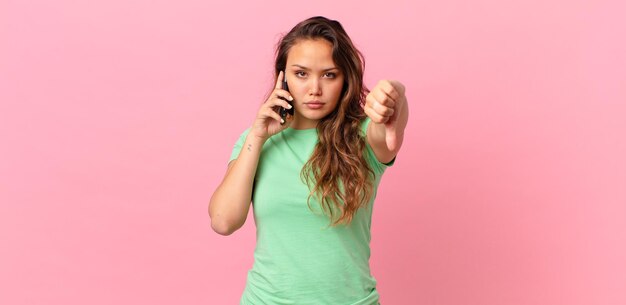 Young pretty woman feeling cross,showing thumbs down and holding a smart phone