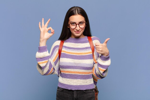 Giovane donna graziosa che si sente felice, stupita, soddisfatta e sorpresa, mostrando gesti bene e pollice in alto, sorridendo. concetto di studente