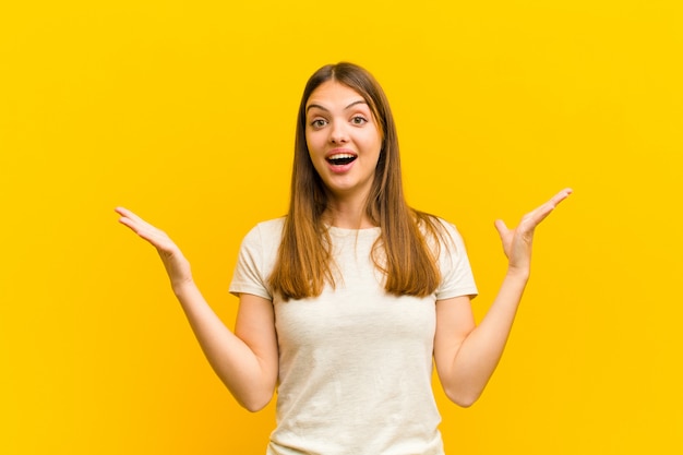 Young pretty woman feeling happy, excited, surprised or shocked, smiling and astonished at something unbelievable against orange background