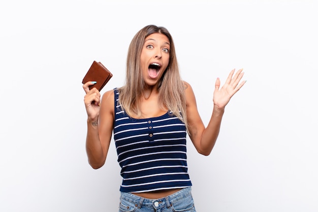 Young pretty woman feeling happy, excited, surprised or shocked, smiling and astonished at something unbelievable with a leather wallet.