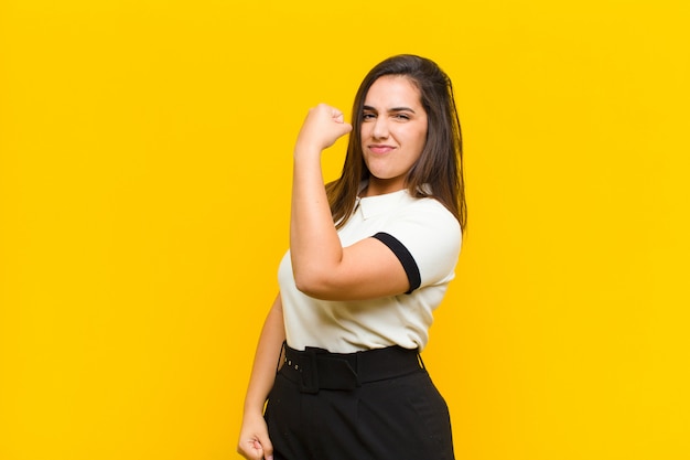 Young pretty woman feeling happy, satisfied and powerful, flexing fit and muscular biceps, looking strong after the gym isolated against orange wall