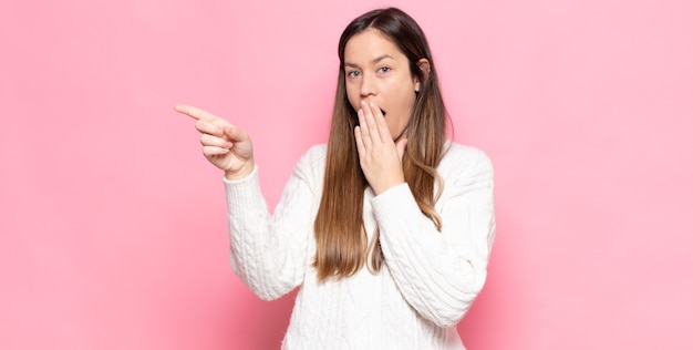 Young pretty woman feeling happy, shocked and surprised, covering mouth with hand and pointing to lateral copy space