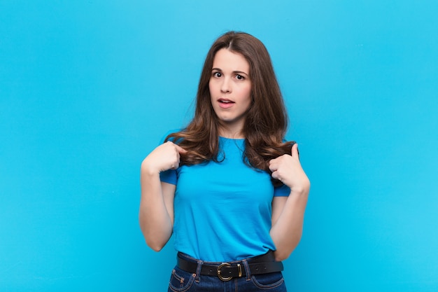 Young pretty woman feeling happy, surprised and proud, pointing to self with an excited, amazed look against blue wall