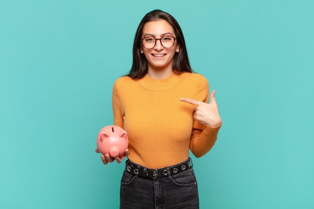 Young pretty woman feeling happy, surprised and proud, pointing to self with an excited, amazed look. piggy bank concept