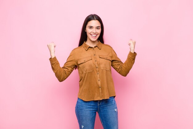 Young pretty woman feeling happy, surprised and proud, shouting and celebrating success with a big smile against pink wall