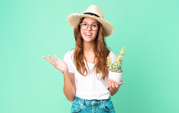 Young pretty woman feeling happy, surprised realizing a solution or idea with a straw hat and holding a cactus