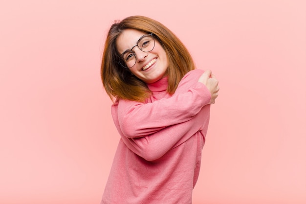 Young pretty woman feeling in love, smiling, cuddling and hugging self, staying single, being selfish and egocentric over pink wall