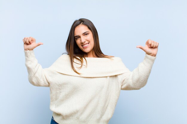 Young pretty woman feeling proud, arrogant and confident, looking satisfied and successful, pointing to self against blue wall