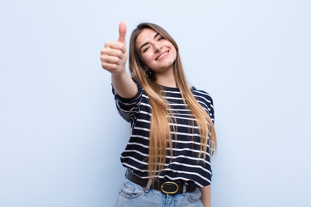 Young pretty woman feeling proud, carefree, confident and happy, smiling positively with thumbs up against soft blue wall