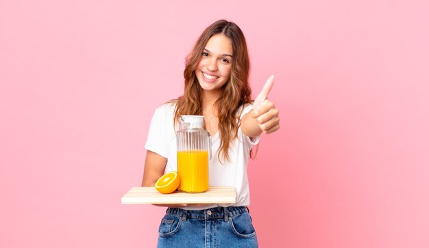 Young pretty woman feeling proud,smiling positively with thumbs up and holding a tray with an orange juice