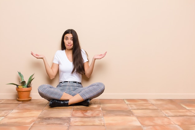 Young pretty woman feeling puzzled and confused, doubting, weighting or choosing different options with funny expression sitting on a floor