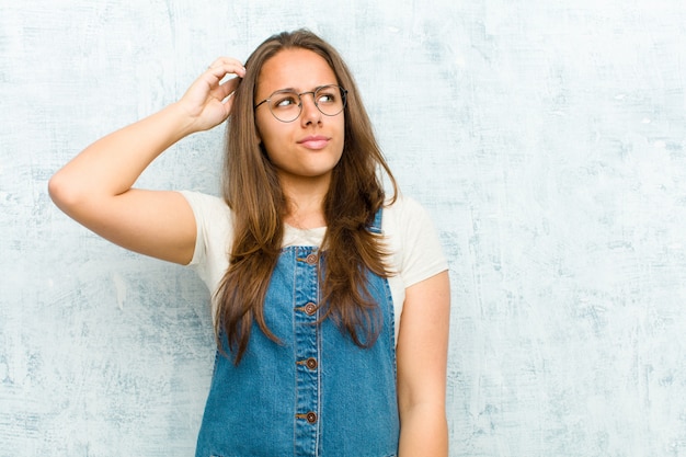 Young pretty woman feeling puzzled and confused, scratching head and looking to the side against grunge wall