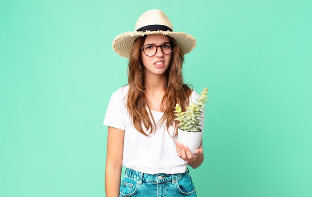Young pretty woman feeling puzzled and confused with a straw hat and holding a cactus