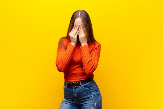 Young pretty woman feeling sad, frustrated, nervous and depressed, covering face with both hands, crying against orange