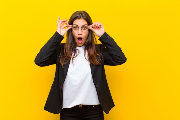 Young  pretty woman feeling shocked, amazed and surprised, holding glasses with astonished, disbelieving look on orange wall