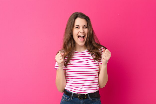 Young pretty woman feeling shocked, excited and happy, laughing and celebrating success, saying wow! against pink wall