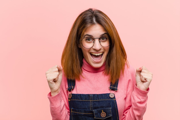 Young pretty woman feeling shocked, excited and happy, laughing and celebrating success, saying wow! against pink wall