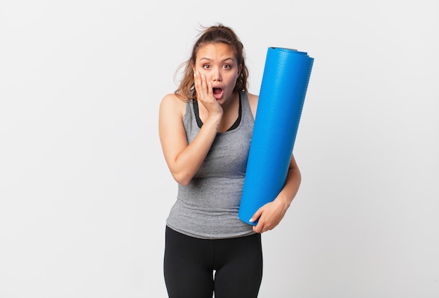 Young pretty woman feeling shocked and scared and holding a yoga mat