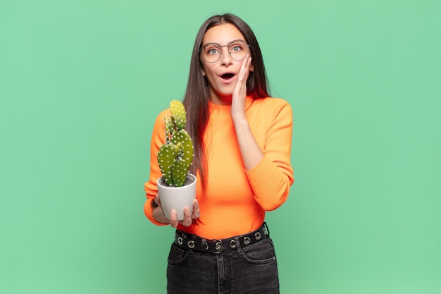 Photo young pretty woman feeling shocked and scared, looking terrified with open mouth and hands on cheeks. cactus concept