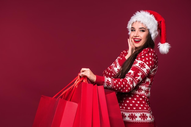 Young pretty woman or girl with long beautiful hair in Santa hat and New Year's dress holding red package on red studio background