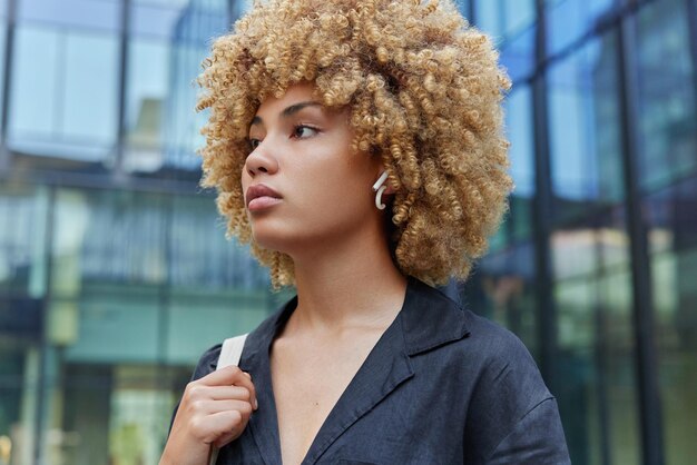 Young pretty woman has curly hair wears black shirt\
concentrated into distance with thoughtful expression strolls\
against modern glass urban building spends free time in city centre\
lifestyle concept