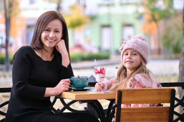 Young pretty woman having good time with her child daughter sitting at street cafe with hot drinks on sunny autumn day Happiness in family relations concept