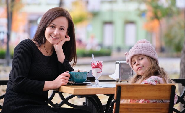 Young pretty woman having good time with her child daughter sitting at street cafe with hot drinks on sunny autumn day Happiness in family relations concept