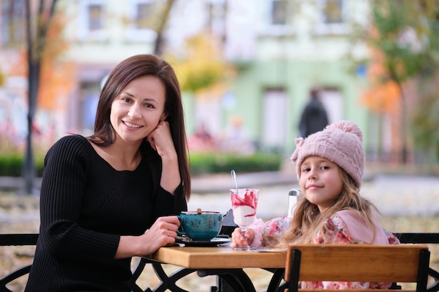 Young pretty woman having good time with her child daughter sitting at street cafe with hot drinks on sunny autumn day Happiness in family relations concept