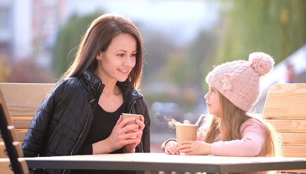 Young pretty woman having good time with her child daughter sitting at street cafe with hot drinks on sunny autumn day Happiness in family relations concept