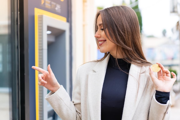 Giovane donna graziosa che tiene un bitcoin all'aperto che indica il lato per presentare un prodotto