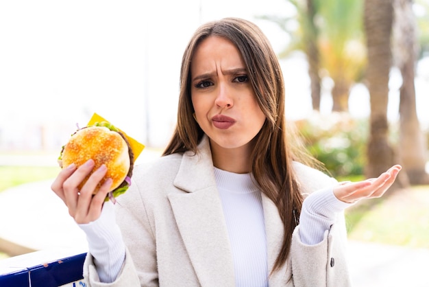 Young pretty woman holding a burger at outdoors making doubts gesture while lifting the shoulders