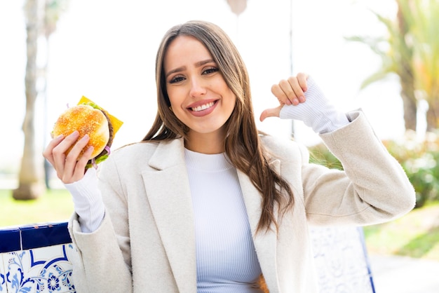 Young pretty woman holding a burger at outdoors proud and selfsatisfied