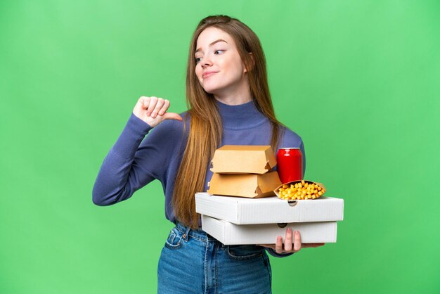 Young pretty woman holding pizzas and burgers over isolated chroma key background proud and selfsatisfied