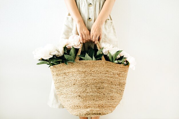 Young pretty woman holding straw bag with white peony flowers on white surface