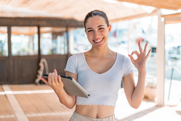 young pretty woman holding a tablet at outdoors showing ok sign with fingers