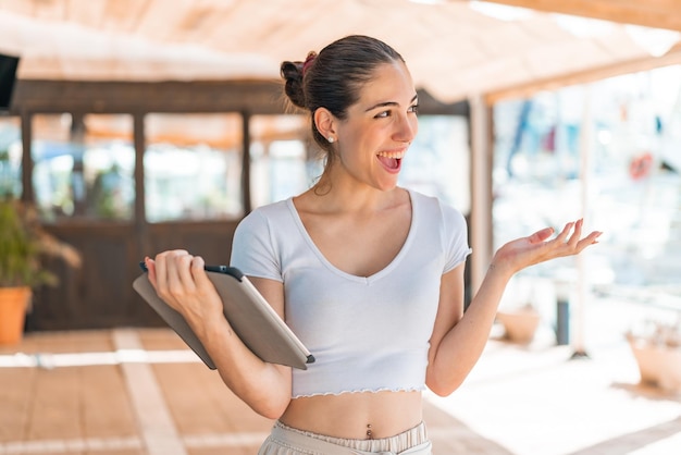 Young pretty woman holding a tablet at outdoors with surprise facial expression