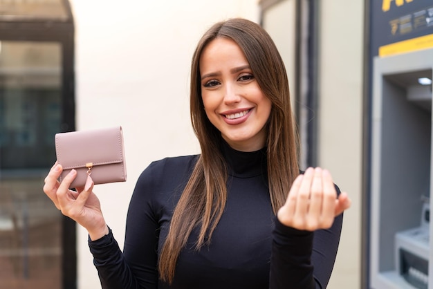 Giovane bella donna che tiene un portafoglio all'aperto che invita a venire con la mano felice che tu sia venuto