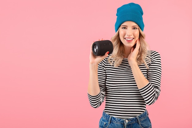 Young pretty woman holding wireless speaker listening to music wearing striped shirt and blue hat smiling happy positive mood posing on pink background isolated